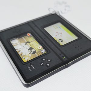 Op de afbeelding: Een zwarte en grijze Nintendo DS handheld-spelconsole met een wit en geel scherm waarop een hond in bad te zien is. De console heeft knoppen met de labels "Start", "Select", "Y", "X", "B" en "A".