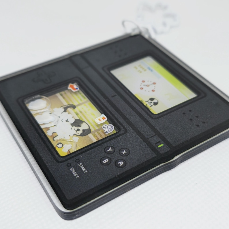 Op de afbeelding: Een zwarte en grijze Nintendo DS handheld-spelconsole met een wit en geel scherm waarop een hond in bad te zien is. De console heeft knoppen met de labels "Start", "Select", "Y", "X", "B" en "A".