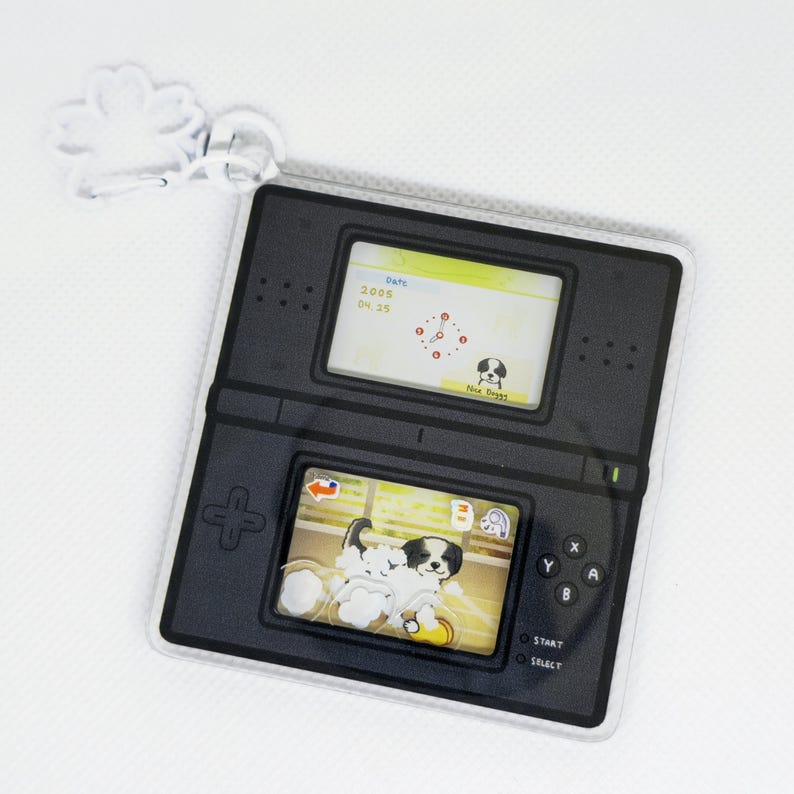 Op de afbeelding: Een zwart-witte sleutelhanger in de vorm van een Nintendo DS handheld-spelconsole. Het scherm toont een spel met een hondenpersonage. De sleutelhanger heeft een zilveren metalen lus om aan een tas of sleutels te bevestigen.