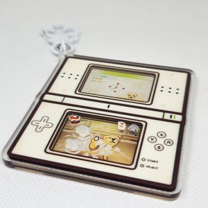 Op de afbeelding: Een witte en bruine Nintendo DS-sleutelhanger met een cartoonhond op het scherm. Het scherm toont de datum "2008 04 23" en een klok. De knoppen zijn gelabeld met "Start" en "Select".