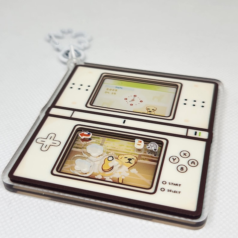 Op de afbeelding: Een witte en bruine Nintendo DS-sleutelhanger met een cartoonhond op het scherm. Het scherm toont de datum "2008 04 23" en een klok. De knoppen zijn gelabeld met "Start" en "Select".