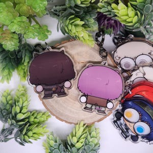 Op de afbeelding: Acryl sleutelhangers met cartoonfiguren met pluizig haar en gedetailleerde outfits. E&eacute;n sleutelhanger heeft bruin haar, de andere heeft paars haar. De sleutelhangers liggen op een houten schijf, omringd door groen.