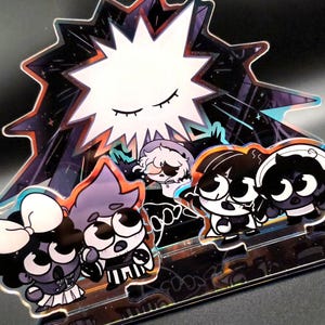 Puede incluir: Una pieza decorativa de acrílico con personajes de dibujos animados con ojos grandes y un diseño de explosión estelar. Los personajes están en varias poses, con una estrella blanca en el fondo. La paleta de colores incluye negro, blanco, morado y naranja.