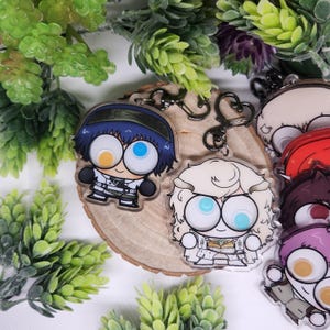 Op de afbeelding: Verschillende sleutelhangers met anime-personages, elk met grote ogen en unieke outfits. E&eacute;n sleutelhanger toont een personage met blauw haar en een uniform in militaire stijl. De sleutelhangers zijn bevestigd aan zilverkleurige sluitingen en worden gepresenteerd op een houten schijf.