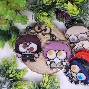 Op de afbeelding: Sleutelhangers met cartoonpersonages met grote ogen en unieke kapsels. E&eacute;n sleutelhanger heeft een personage met bruin haar en een gestreept outfit, een andere heeft roze haar en een geruit jasje. De sleutelhangers zijn bevestigd aan zilveren sleutelringen.