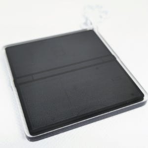 Op de afbeelding: Een zwart-witte sleutelhanger in de vorm van een draagbare gameconsole. De sleutelhanger is gemaakt van helder acryl en heeft een zwart-witte afbeelding van een gameconsole erop.