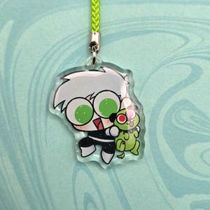 May include: A clear acrylic charm featuring a cartoon character with white hair, large green eyes, and a black and white outfit, holding a green creature with a red eye. The charm is attached to a green lanyard.