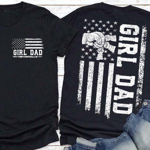 May include: A black short-sleeve t-shirt featuring a white graphic of an American flag and the text GIRL DAD on the front left chest. The back of the shirt displays a larger version of the flag, a fist bump graphic, and the text GIRL DAD.