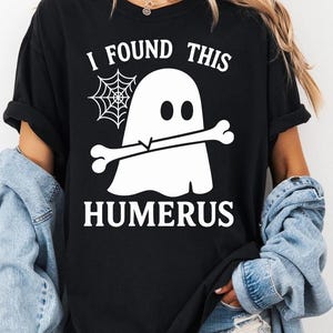 May include: Black t-shirt with a white ghost graphic holding a bone, a spiderweb, and the text "I FOUND THIS HUMERUS". The shirt is a Halloween-themed design.