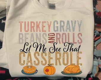 May include: White sweatshirt with the text "TURKEY GRAVY BEANS AND ROLLS Let Me See That CASSEROLE". The design includes illustrations of a pie, pumpkin, and roasted turkey. A festive garment for Thanksgiving.
