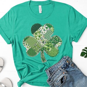 May include: A teal green t-shirt featuring a large shamrock design. The shamrock is composed of various green patterned fabrics, including floral, polka dot, and paisley prints. The shirt is paired with blue denim jeans.