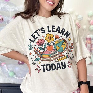 May include: Cream-colored t-shirt with the text "LET'S LEARN TODAY" in a whimsical font. The design includes a globe, books, flowers, and a pencil. The shirt is a comfortable, casual style.
