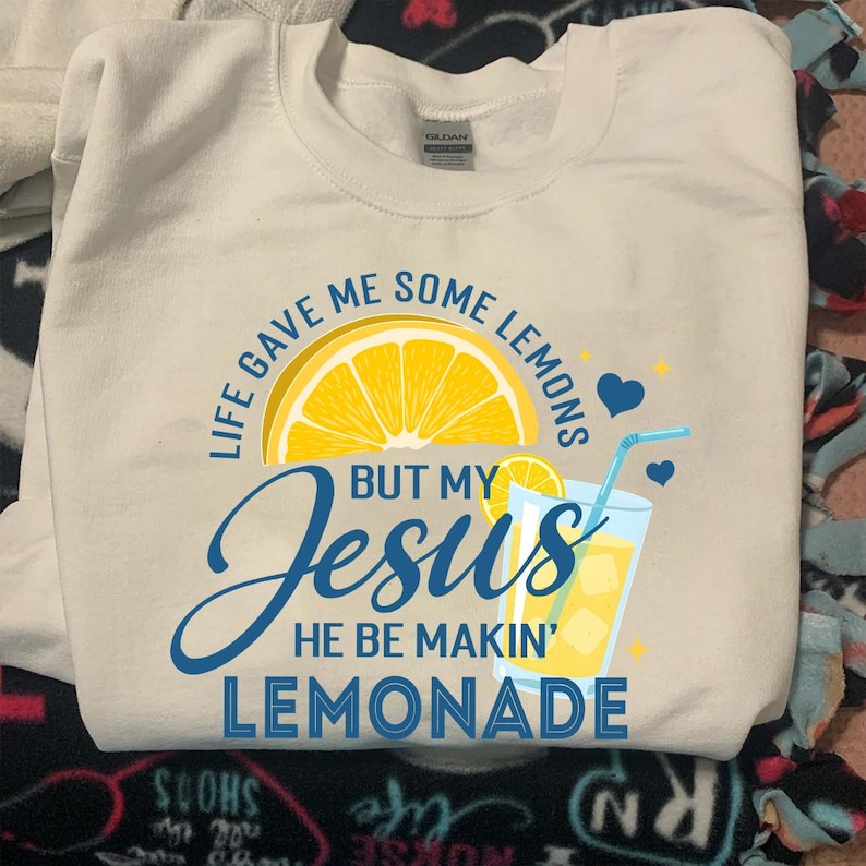May include: A white Gildan sweatshirt with a graphic design. The design features a yellow lemon slice, a glass of lemonade, and the text "LIFE GAVE ME SOME LEMONS BUT MY Jesus HE BE MAKIN' LEMONADE" in blue and yellow.