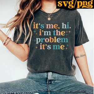 May include: A dark gray t-shirt with a colorful graphic that reads "it's me, hi. i'm the problem it's me". The graphic features stars and a distressed look.