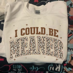 May include: Cream-colored sweatshirt with the text "I COULD BE MEANER" in brown and leopard print. The sweatshirt has a crew neck and long sleeves. The text is surrounded by small starburst designs.