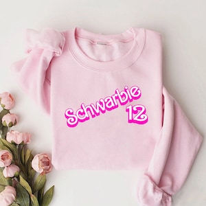 May include: Pink sweatshirt with a pink graphic that says "Schwarbie 12" in a retro font.