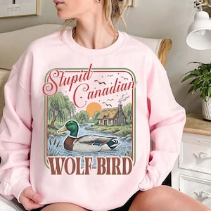 Könnte beinhalten: Hellrosa Sweatshirt mit einem Grafikdesign, das eine Ente in einem Fluss, eine Hütte und den Text "Stupid Canadian WOLF-BIRD" zeigt. Das Design hat eine Vintage- und Outdoor-Ästhetik.