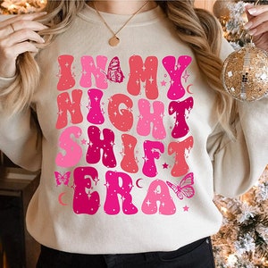 Peut inclure: Un sweat-shirt crème avec le texte "In My Night Shift Era" en lettres roses et rouges avec des accents de papillon et d'étoile.