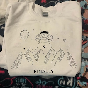 Op de afbeelding: Witte sweatshirt met een zwart-witte afbeelding van een UFO die een persoon van een bergketen ontvoert, met een maan en sterren op de achtergrond. Het woord "FINALLY" staat eronder gedrukt.