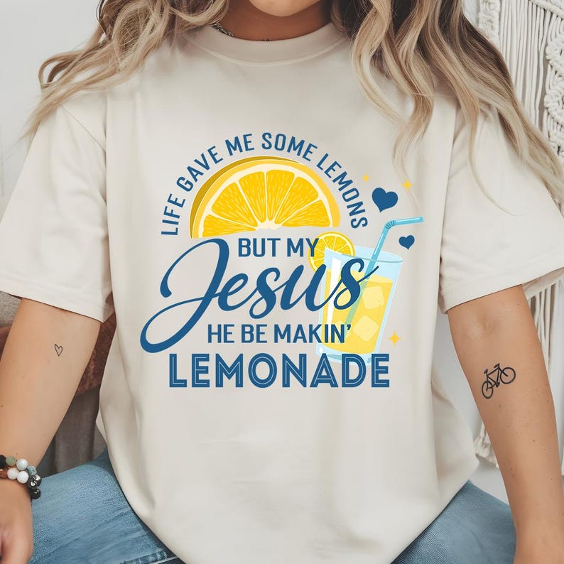 May include: Cream-colored t-shirt with a graphic design featuring a lemon slice, a glass of lemonade, and the text "Life gave me some lemons but my Jesus he be makin' lemonade." The design includes blue hearts and stars.