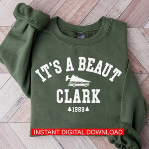 May include: A green sweatshirt with white text that reads "It's a beaut Clark 1989" with a graphic of a car with a Christmas tree on top.