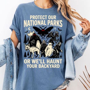 Puede incluir: Camiseta azul con el texto "PROTECT OUR NATIONAL PARKS OR WE'LL HAUNT YOUR BACKYARD". El gráfico presenta una polilla negra, fantasmas, un dragón y otras criaturas espeluznantes. La camiseta tiene las mangas enrolladas.