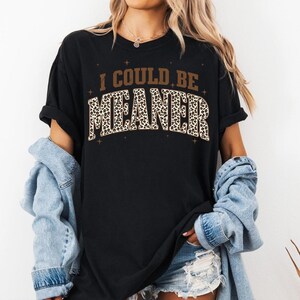 Puede incluir: Camiseta negra con el texto "I COULD BE MEANER" en letras marrones y estampadas de leopardo. La camiseta tiene mangas cortas y un corte holgado. Parte de una colecci&oacute;n de ropa.