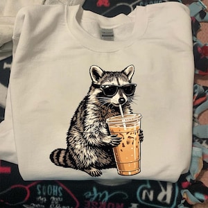 May include: White sweatshirt featuring a graphic of a raccoon wearing sunglasses and drinking iced coffee. The raccoon is black, white, and gray. The iced coffee is in a clear plastic cup with a straw.
