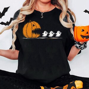 May include: Black t-shirt with a Halloween design. The graphic shows an orange pumpkin 'eating' three small white ghosts. A small orange pumpkin-shaped mug is held in the hand. The image has a Halloween theme.