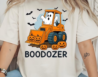 May include: Cream-colored t-shirt featuring a Halloween-themed graphic. The design includes an orange bulldozer with a ghost driver, jack-o'-lanterns, and bats. The word "BOODOZER" is printed in black block letters below the bulldozer.