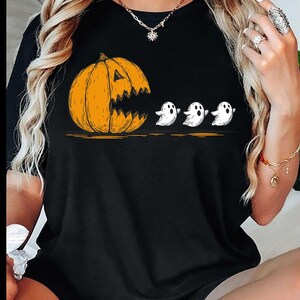 Puede incluir: Camiseta negra con un diseño de Halloween. Una calabaza naranja con boca dentada persigue a tres fantasmas blancos. Una ilustración divertida para la temporada de otoño.
