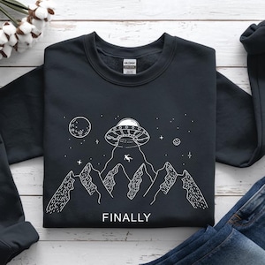 Op de afbeelding: Zwart sweatshirt met een witte afbeelding van een UFO die een persoon ontvoert boven een bergketen, met een maan en sterren op de achtergrond. Het woord "FINALLY" staat eronder.