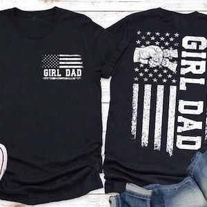 May include: Black t-shirts with the text "GIRL DAD" and an American flag design. One shirt has the flag on the front pocket, the other on the back with a fist bump graphic. White sneakers and blue jeans are also visible.