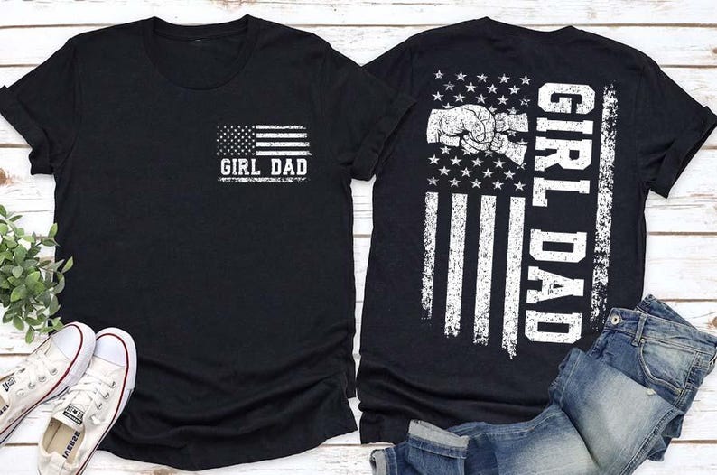 Puede incluir: Dos camisetas negras con el texto "GIRL DAD" y un dise&ntilde;o de bandera estadounidense. Una tiene la bandera en el bolsillo, la otra en la espalda con un gr&aacute;fico de choque de pu&ntilde;os. Tambi&eacute;n se ven zapatillas blancas y vaqueros azules.