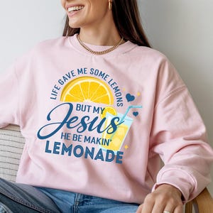 May include: Pink sweatshirt with a graphic that says "Life gave me some lemons but my Jesus he be makin' lemonade." The design includes a yellow lemon slice, a glass of lemonade, and blue hearts.