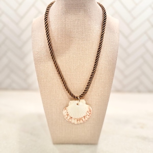 May include: A seashell pendant necklace displayed on a mannequin. The necklace features a brown rope-like cord and a white seashell pendant with reddish-brown accents. The pendant is attached with a gold-colored bail.