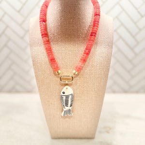 May include: A coral-colored beaded necklace with a gold clasp and a white fish pendant with black detailing. The necklace is displayed on a beige mannequin. The fish pendant has a minimalist design.