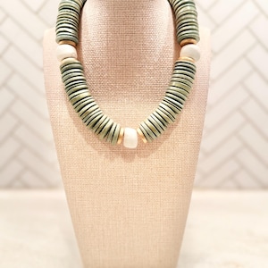May include: A chunky necklace with a mix of green, white, and gold-toned beads. The necklace is displayed on a beige jewelry bust. The beads are round and flat, creating a layered effect. The necklace is a statement piece.