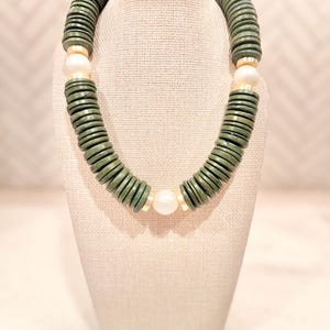 May include: A chunky necklace featuring olive green disc-shaped beads and three large, off-white, round beads. The necklace is displayed on a neutral-colored bust, showcasing its design and texture. The necklace is a statement piece.