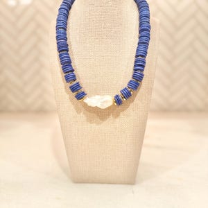 May include: A blue beaded necklace with a white stone pendant and gold accents. The necklace is displayed on a beige jewelry stand. The beads are round and evenly spaced, creating a bold, eye-catching accessory.