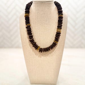 May include: A dark brown beaded necklace with a mix of round and flat beads. The necklace is displayed on a beige jewelry stand. The necklace has a mix of dark brown and light brown beads.