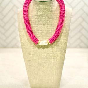 May include: A vibrant pink necklace displayed on a beige jewelry bust. The necklace features a series of round, flat pink beads. A large, off-white, irregular-shaped bead is centered, with gold-colored accents. The necklace is a bold statement piece.