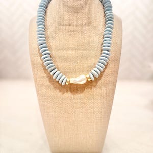 May include: A necklace featuring a series of light blue, disc-shaped beads arranged in a circular pattern. At the center, a single white, irregular-shaped pearl is flanked by two small, cylindrical gold-toned metal spacers, completing the design.