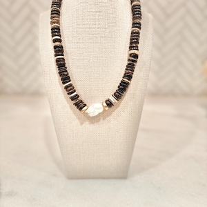 May include: A handcrafted necklace featuring dark brown, light brown, and white wooden beads. The necklace has a central white stone pendant and gold-colored accents. The beads are strung together to create a stylish accessory.