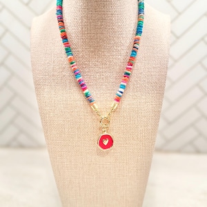 May include: A colorful beaded necklace with a gold clasp and a red pendant. The pendant features a small white heart. The necklace is displayed on a beige mannequin.