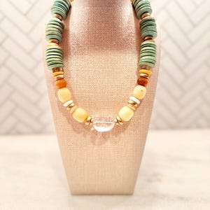 May include: A necklace with a variety of beads in shades of green, yellow, and orange. The necklace features large, round, green beads, smaller yellow and orange beads, and a clear, round pendant. The beads are strung together with gold-colored spacers.