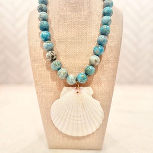 May include: A necklace featuring a strand of round, variegated blue and teal stone beads. A large, white, fan-shaped seashell pendant is attached to the center of the bead strand with a small gold-toned metal jump ring, creating a coastal-inspired piece.