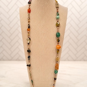 May include: A long necklace featuring a variety of multicolored beads in different shapes and sizes, including round, oval, and rectangular forms. The beads are made of various materials in shades of orange, green, brown, and clear, interspersed with small wooden spacer beads along the entire length of the strand.