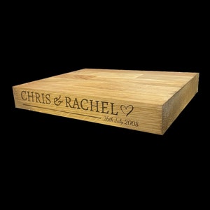 Op de afbeelding: Een lichtgekleurd houten blok met gegraveerde tekst. De tekst luidt "CHRIS & RACHEL" boven een hartsymbool en de datum "26th July 2008". Het blok is rechthoekig en lijkt een decoratief item te zijn.