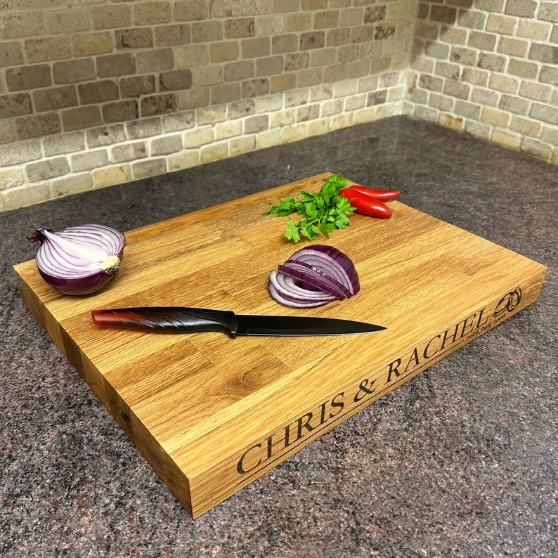 Op de afbeelding: Een houten snijplank met de namen "CHRIS & RACHEL" gegraveerd. Er liggen een zwart mes, rode uien, peterselie en rode pepers op. De plank is gemaakt van lichtbruin hout.